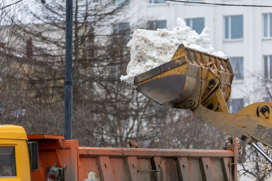 Excavator clearing snow into dump truck on city street during winter.